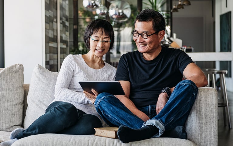 Asian couple sitting on a sofa at home looking at a tablet.